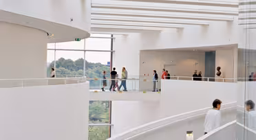 Moderne arkitektonisk interiør med buet trappe og store glasvinduer. Folk går på det øverste niveau med udsigt over grønne områder udenfor. Rummet er lyst med et minimalistisk hvidt design.