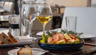 Et bord med en tallerken salat toppet med laks, citronskiver og dild, to glas hvidvin og en lille tallerken brød. Baggrunden har en moderne spisestue med blød belysning.