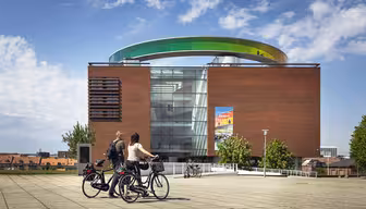 To personer på cykler holder pause foran en moderne murstensbygning med en stor, farverig cirkulær struktur på taget. Himlen er delvist overskyet, og der er en plakat på bygningens glasfacade. Træer og bybilledet er synlige i baggrunden.