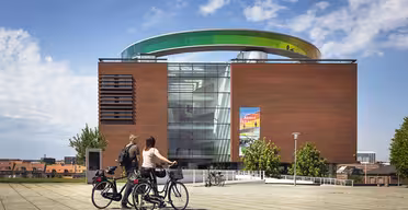To personer på cykler holder pause foran en moderne murstensbygning med en stor, farverig cirkulær struktur på taget. Himlen er delvist overskyet, og der er en plakat på bygningens glasfacade. Træer og bybilledet er synlige i baggrunden.