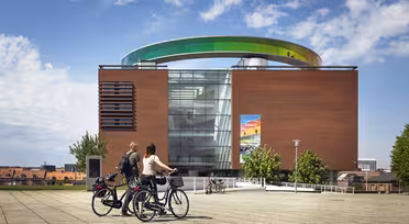 To personer på cykler holder pause foran en moderne murstensbygning med en stor, farverig cirkulær struktur på taget. Himlen er delvist overskyet, og der er en plakat på bygningens glasfacade. Træer og bybilledet er synlige i baggrunden.