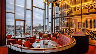 Luksuriøs restaurant med høje vinduer med udsigt over byen. Med røde polstrede kabiner, runde borde til spisning, varm belysning og en dekorativ vinudstillingsvæg. Plys røde tæpper komplementerer den elegante stemning.