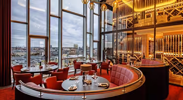 Luksuriøs restaurant med høje vinduer med udsigt over byen. Med røde polstrede kabiner, runde borde til spisning, varm belysning og en dekorativ vinudstillingsvæg. Plys røde tæpper komplementerer den elegante stemning.