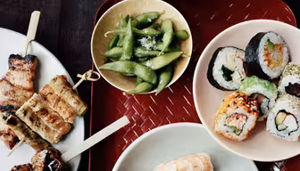 En række japanske retter er kunstfærdigt arrangeret. Der er en tallerken med sushiruller, en tallerken med nigiri sushi, en skål edamame, grillet kød på spyd og en skål ris. Spisepinde hviler i nærheden på den teksturerede røde bakke.