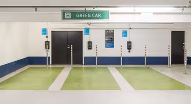Parkeringsplads til elbiler med grønmalede pladser under et "GRØN BIL" skilt. Der er monteret ladestandere på væggen mellem parkeringsbanerne og flere små skilte med instruktioner.